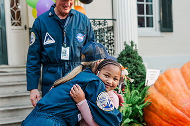 A woman in an astronaut suit bends down to hug a little girl wearing a pink shirt.