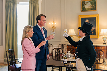 A woman in a pink suit, a man in a blue suit with a pink tie and a man dressed in 18th century period clothing raise glasses for a toast.
